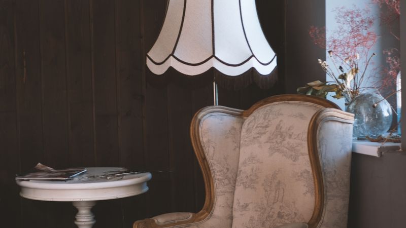 Ohrensessel mit hellem Toile-Bezug unter einer Stehlampe mit weißem Fransenschirm, daneben ein runder weißer Beistelltisch mit Zeitschriften