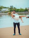 Man in White Shirt and Black Pants Holding and Kissing Woman in White Dress and Shoes with Bouquet of Flowers While Standing on the Beach