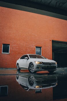 Sleek silver sedan parked by a red brick wall with a wet pavement reflection, showcasing urban style.