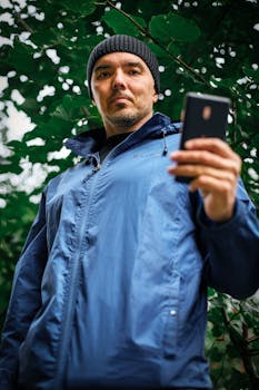A man in a blue jacket holds a smartphone in an outdoor setting with green foliage.