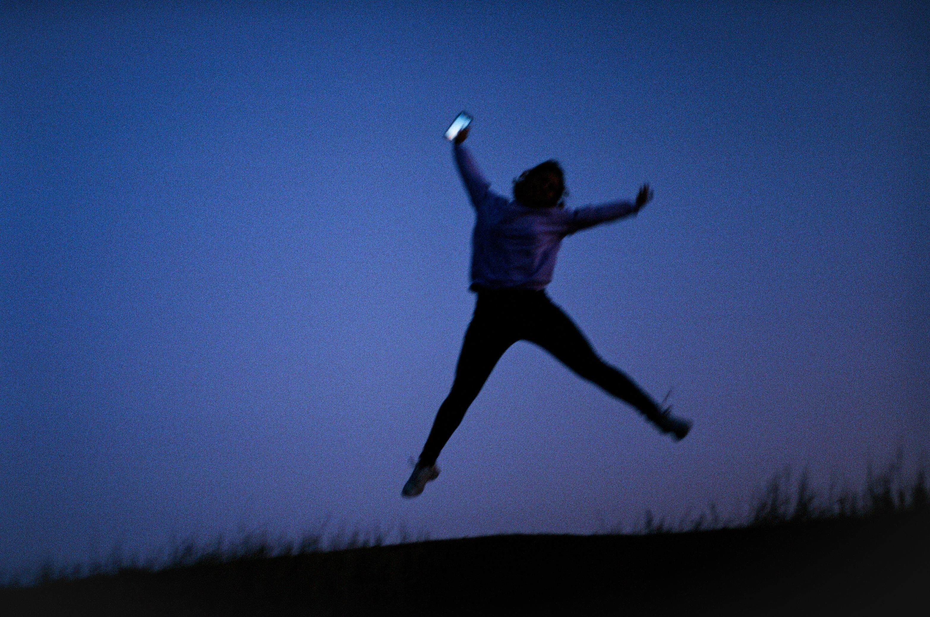 A silhouette of a person energetically jumping with a smartphone against a twilight sky.
