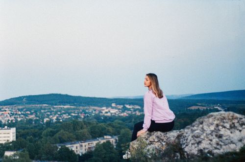 Free Woman relaxing on a cliff overlooking a picturesque countryside, perfect for sightseeing and nature lovers. Stock Photo