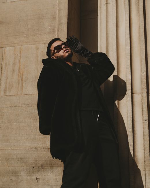 A fashionable man in a black coat poses against a wall in sunlight.
