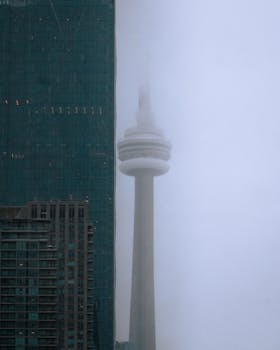 The CN Tower emerges through fog beside modern skyscrapers, creating a dramatic urban scene.