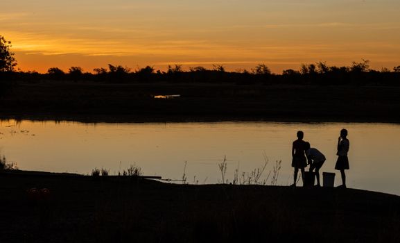 Silhouetted children standing by a tranquil lake during a warm, dramatic sunset.