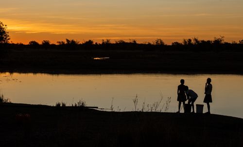 Free Silhouetted children standing by a tranquil lake during a warm, dramatic sunset. Stock Photo