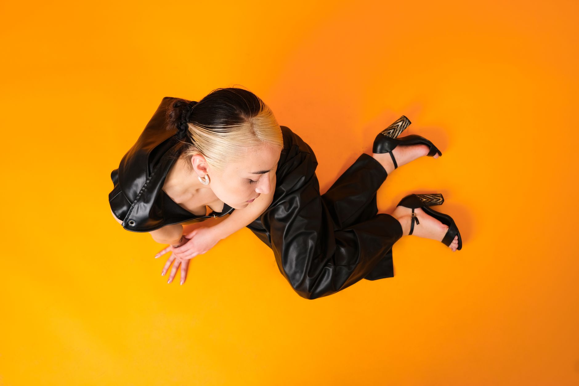 A stylish woman poses in black leather against a vibrant orange backdrop, showcasing contemporary fashion.