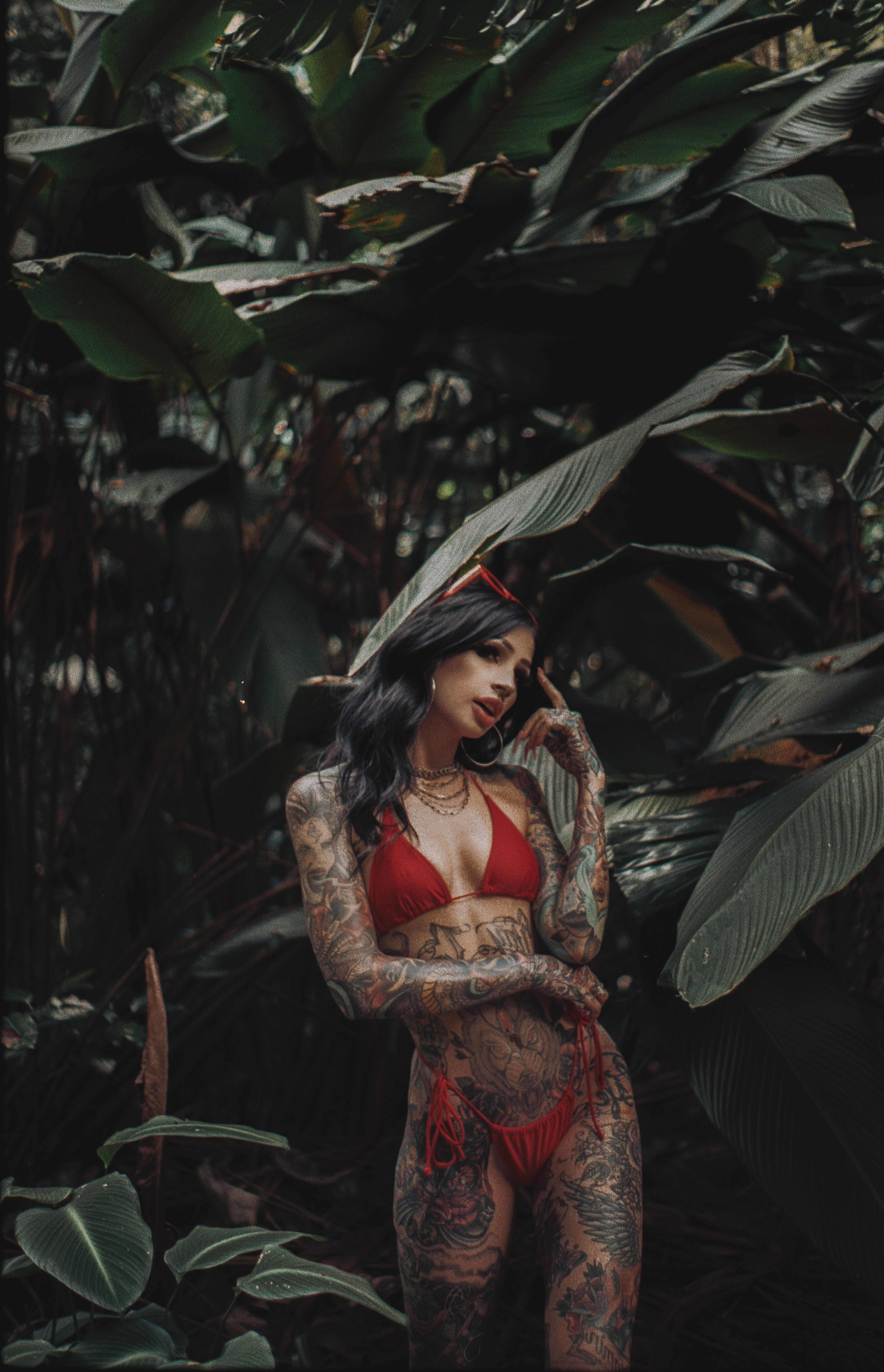 A tattooed woman in a red bikini stands amidst lush tropical foliage, showcasing fashion and nature.