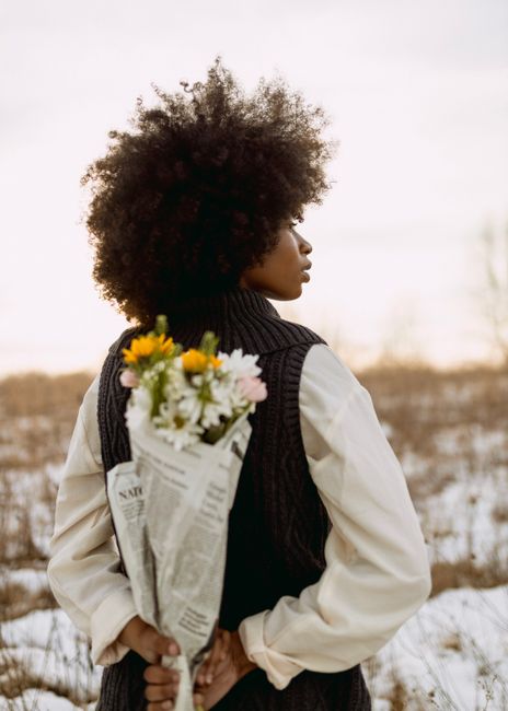 A woman stands in a snowy field holding a flower bouquet behind her back.