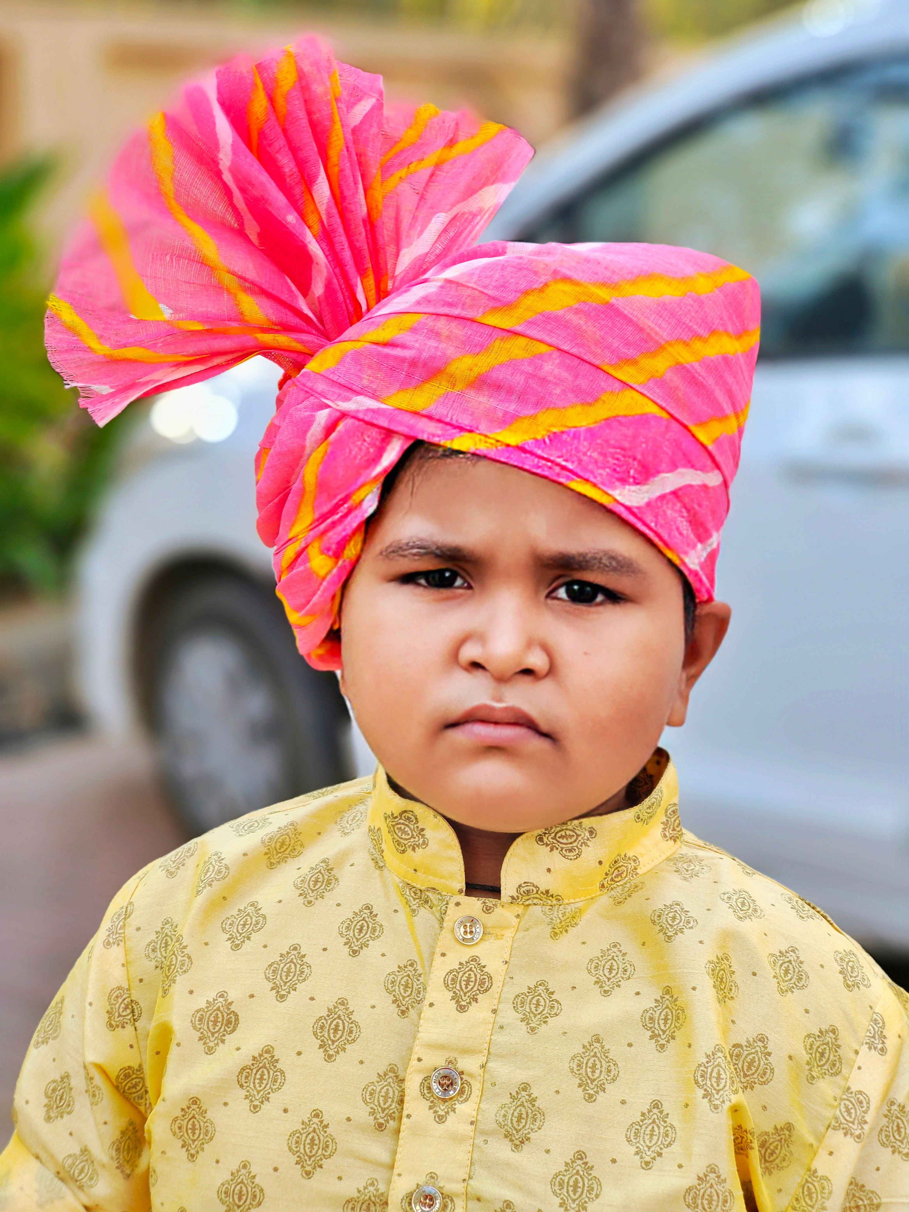 Portrait of a young boy wearing a traditional yellow kurta and vibrant pink turban outdoors.