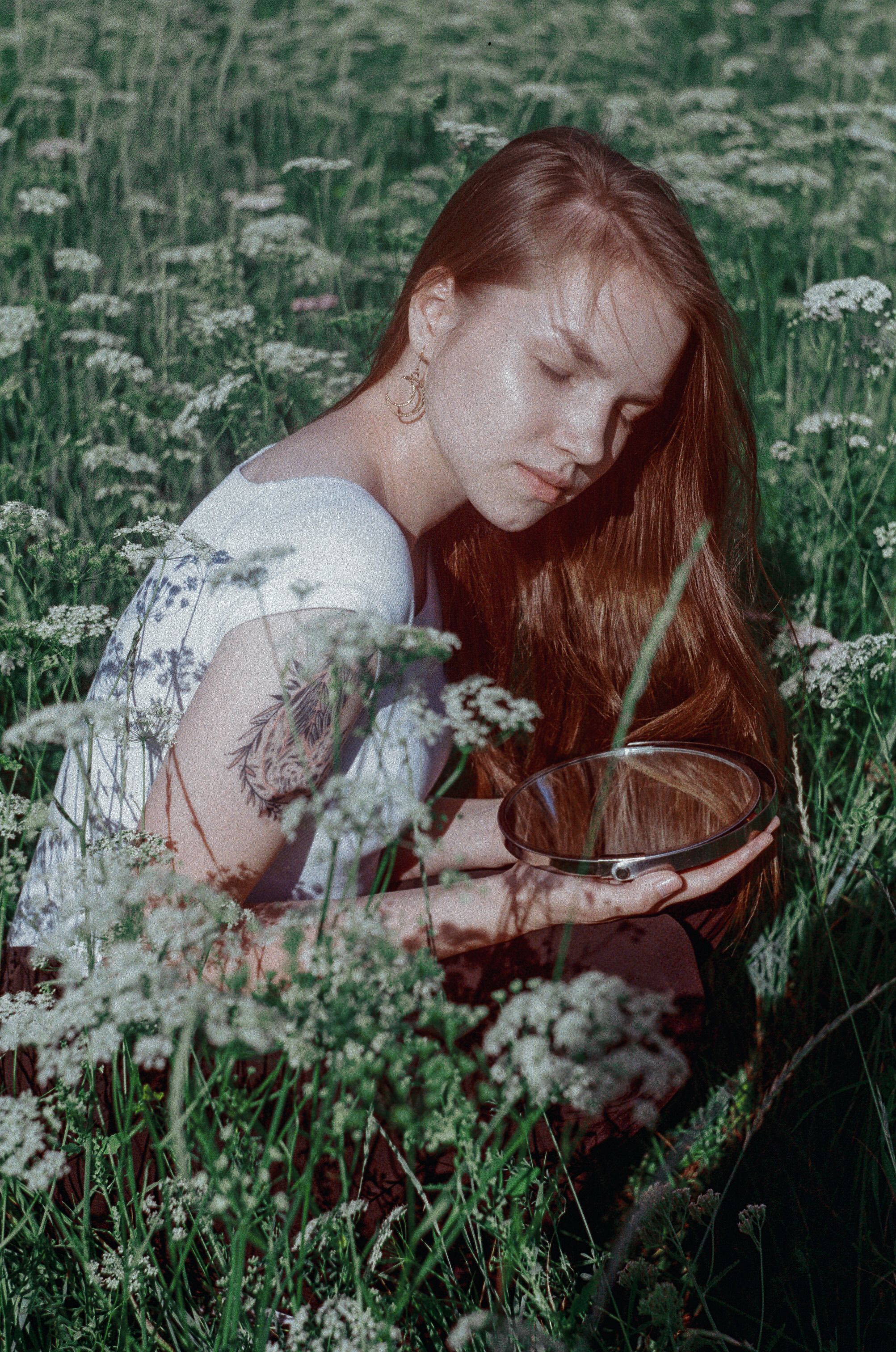 A serene portrait of a young woman with eyes closed, holding a mirror in a lush flower field.