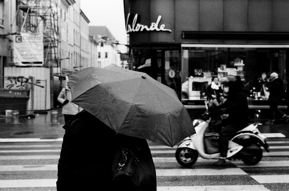 Dynamic city street scene with people walking in the rain and a scooter passing by.