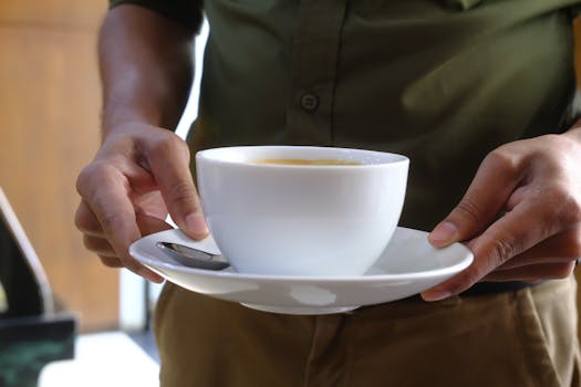 Close-up of hands holding a coffee cup with saucer and teaspoon indoors.
