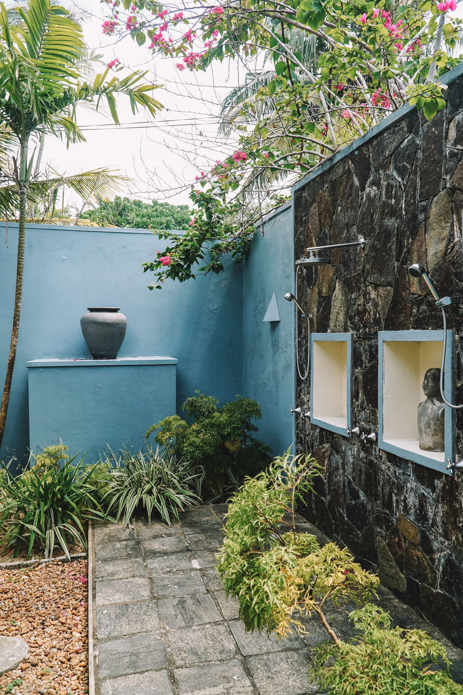 Outdoor garden shower built into a stone wall with tropical planting and an open-air path