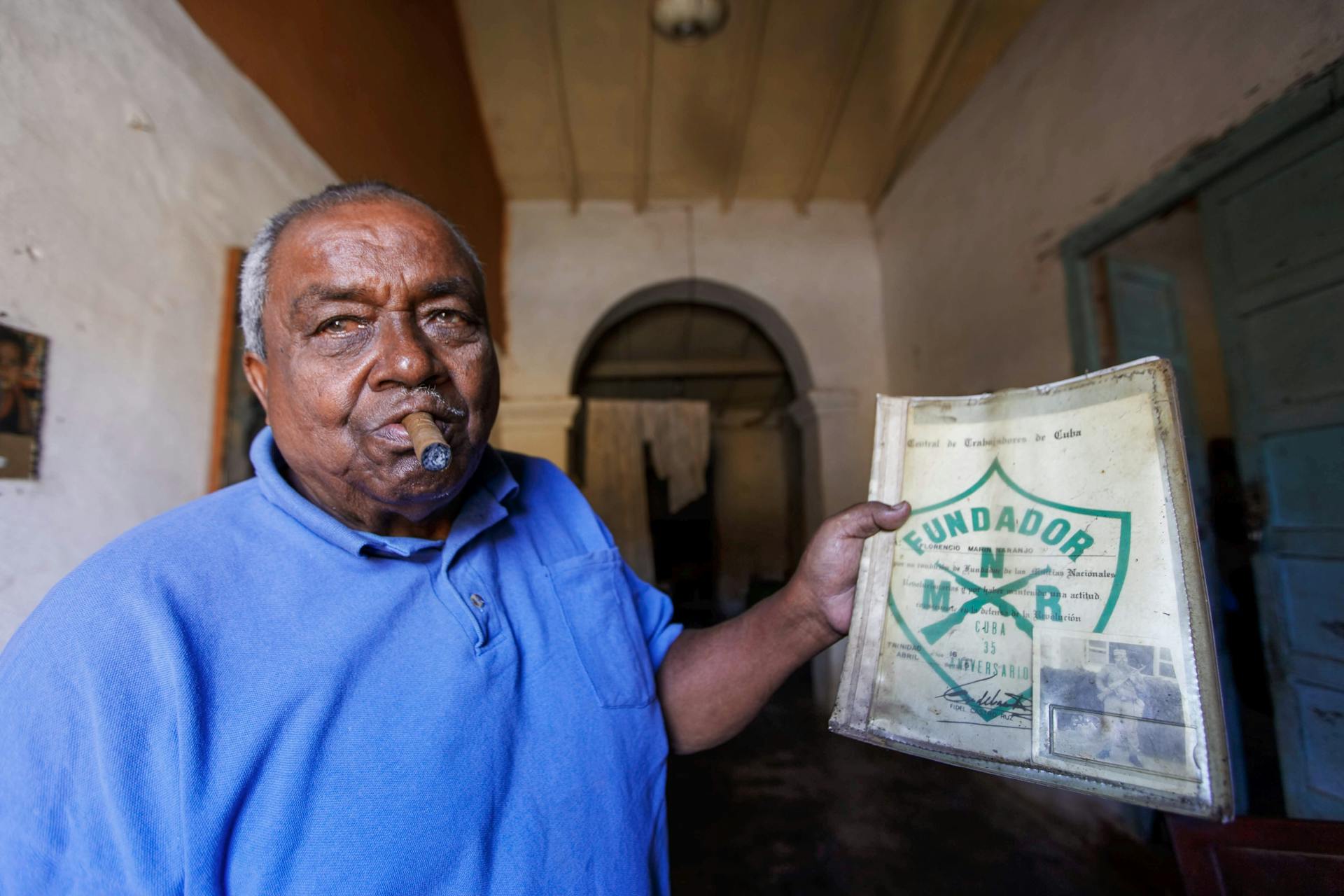 Portrait of an elderly man smoking a cigar and holding a Fundador sign in a rustic indoor setting.