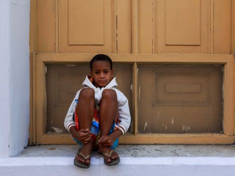 A thoughtful young boy in casual wear sits on a doorstep against a rustic door.
