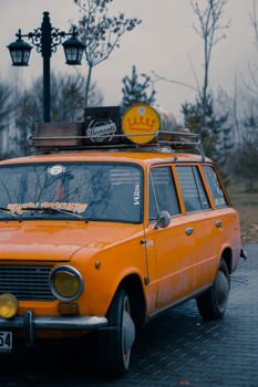 Close-up of a vintage yellow car parked outdoors with fall trees in the background.
