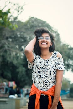 A young woman with glasses smiling outdoors, showcasing fashion with an orange wrap.