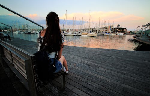 Mulher Em Camisa Preta E Branca Listrada Com Decote Redondo, Sentada No Banco De Madeira Marrom Na Doca Do Navio Durante O Dia