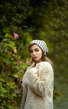 Young woman in a luxurious fur coat and polka dot headscarf holding a smartphone outdoors.