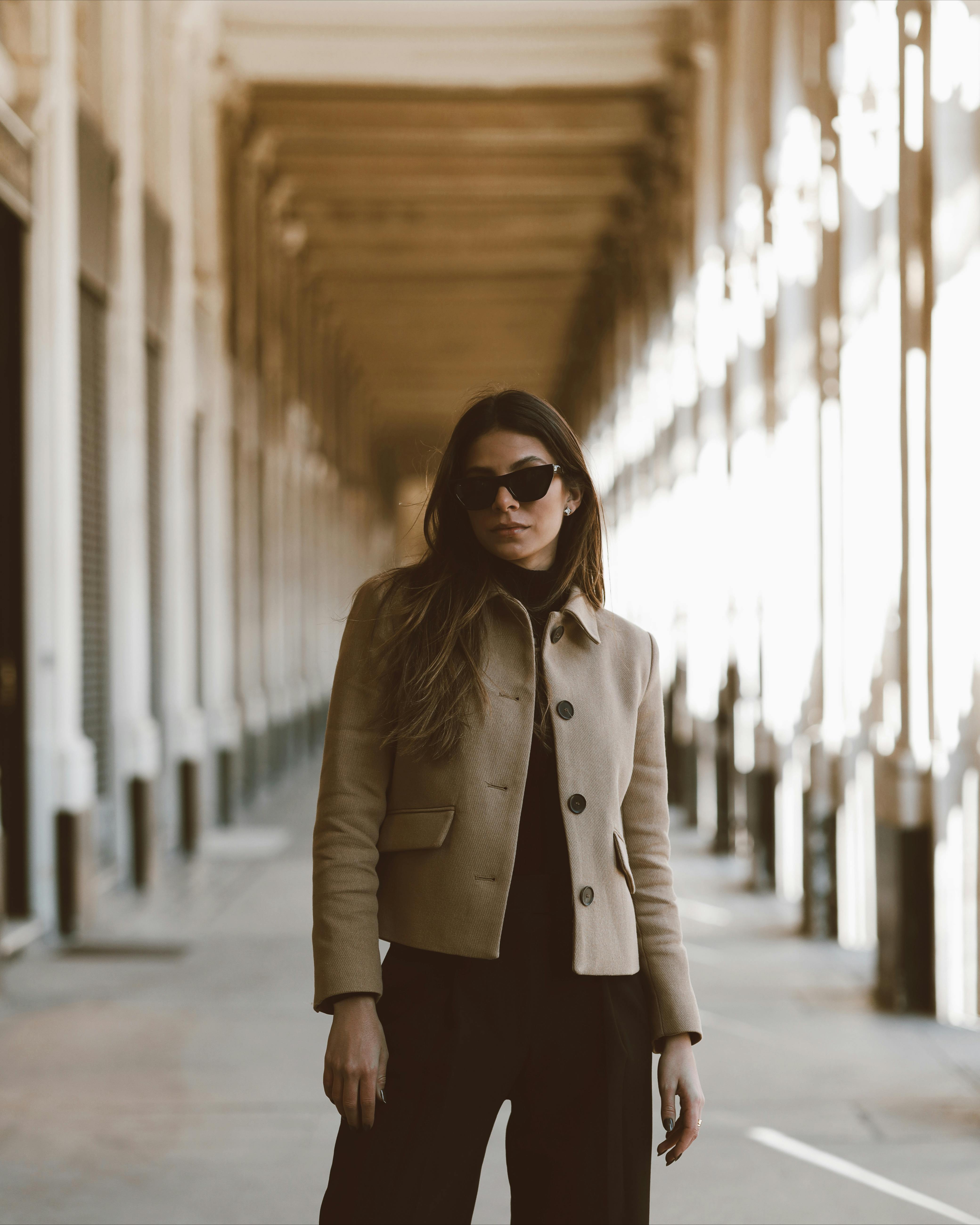 A fashionable woman in a beige coat and sunglasses posing confidently in a classic architectural corridor.