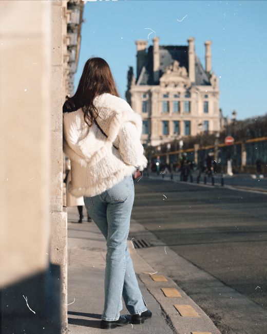 Woman in a white coat leaning against a wall with city architecture in view.