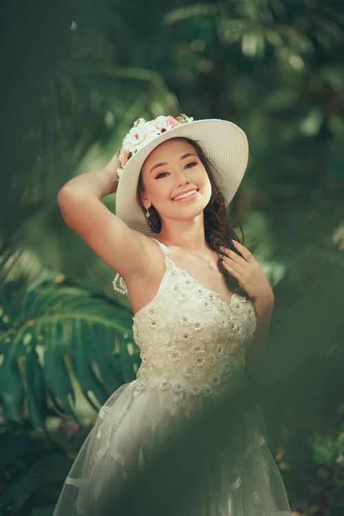 Free Woman in a white dress and floral hat smiling amidst lush greenery in a sunny outdoor setting. Stock Photo