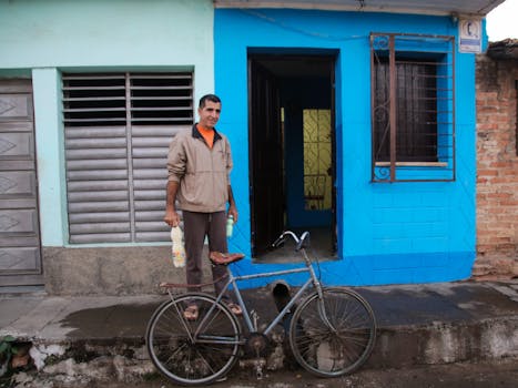 A man stands by his bicycle holding milk bottles in front of a blue and green house.