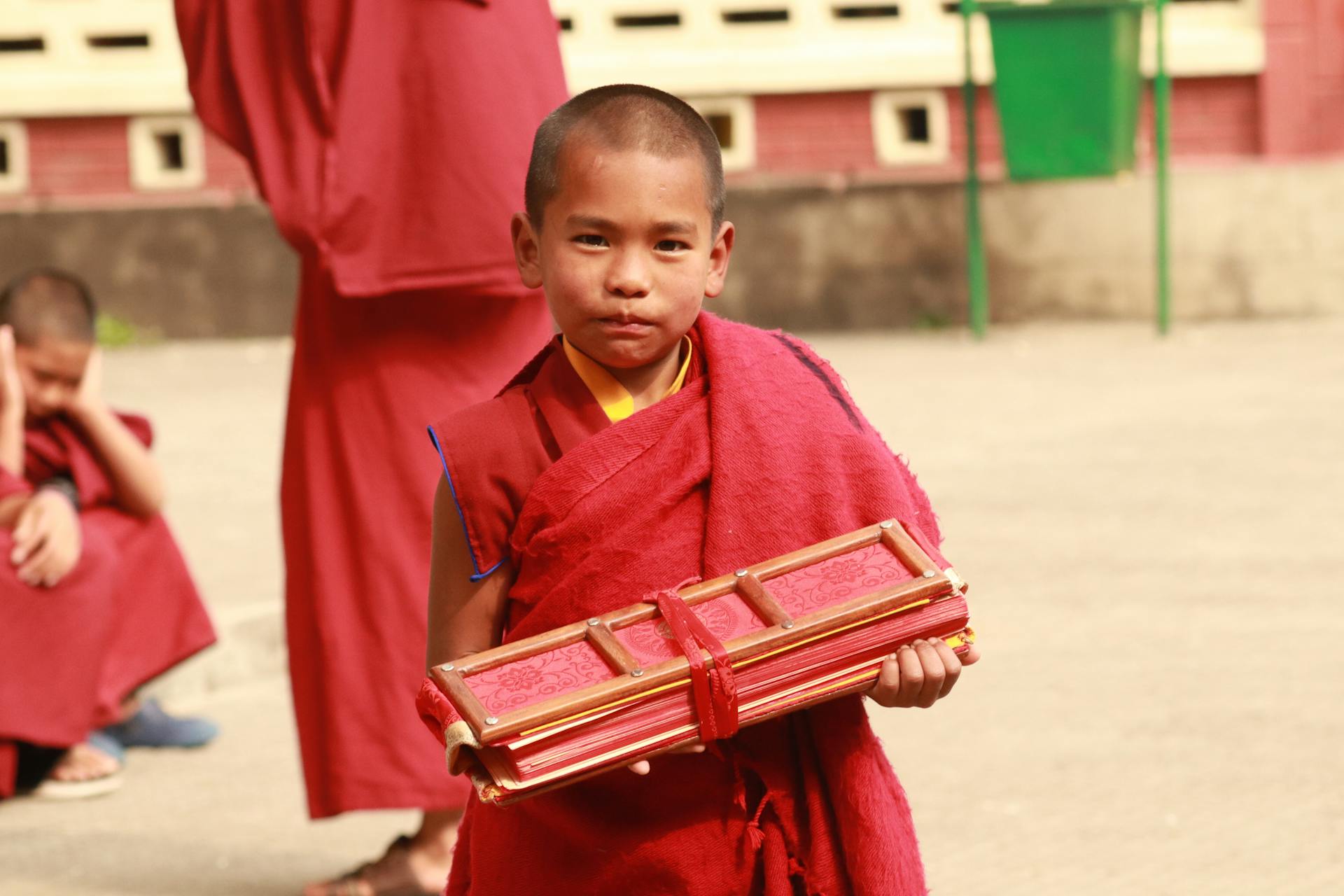 A young Buddhist monk in traditional clothing holding Buddhist scriptures outdoors.