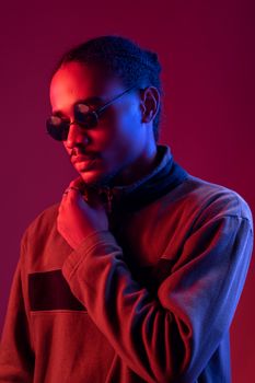 A fashionable man in sunglasses and jacket poses under vibrant red and blue lighting.