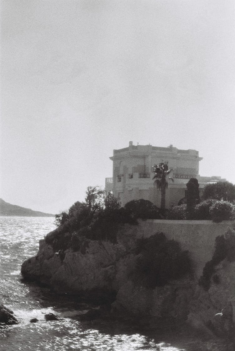 Grayscale Photo Of A Mansion House On Cliff Edge Of Coastal Ocean