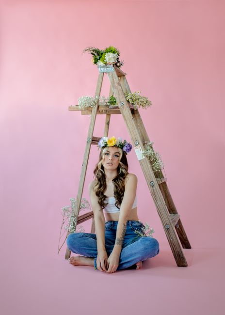 A woman poses with a flower crown, sitting barefoot in jeans against a pink backdrop.