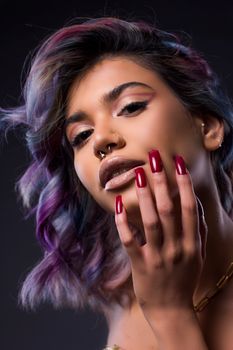 Close-up portrait of a woman with dyed hair, piercing, and vibrant manicure on a dark background.