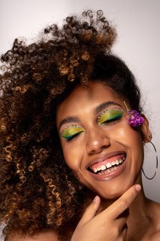 A cheerful woman with curly hair, vibrant green eyeshadow, and floral accents exuding confidence.