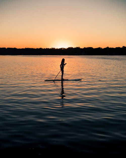 Free A silhouette of a woman paddleboarding at sunset on a tranquil Minnesota lake, backlit by the setting sun. Stock Photo