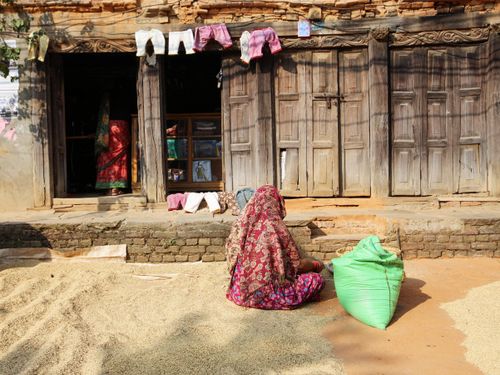 Free A rural woman in an ornate headscarf sits by a rustic house and grain sack. Stock Photo