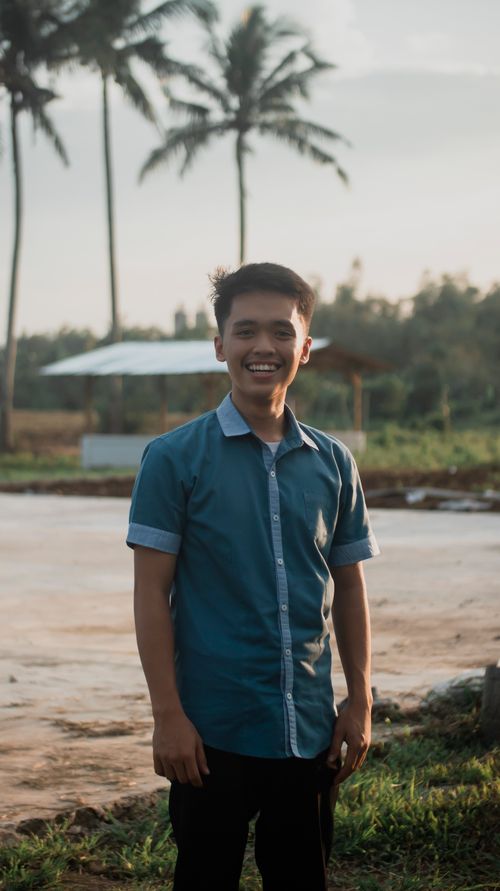 Free Asian man smiling in a blue shirt, standing outdoors with palm trees background. Stock Photo