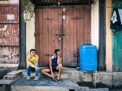 Free Two boys rest outside a rustic wooden gate, with a blue water container beside them. Urban lifestyle scene. Stock Photo