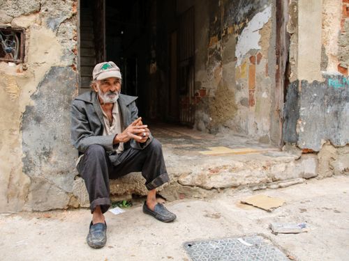 Free Elderly man with a beard sits pensively on a worn sidewalk in front of an aged building. Stock Photo