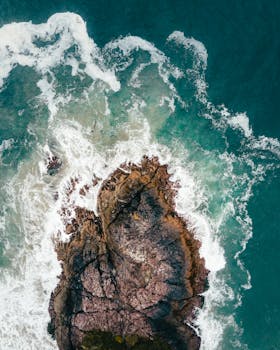 Aerial shot of ocean waves crashing onto rugged rocks in Tofino, Canada, highlighting the dramatic coastal landscape.