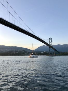 Sailboat travels under Lions Gate Bridge in Vancouver at sunset with mountains in the backdrop.