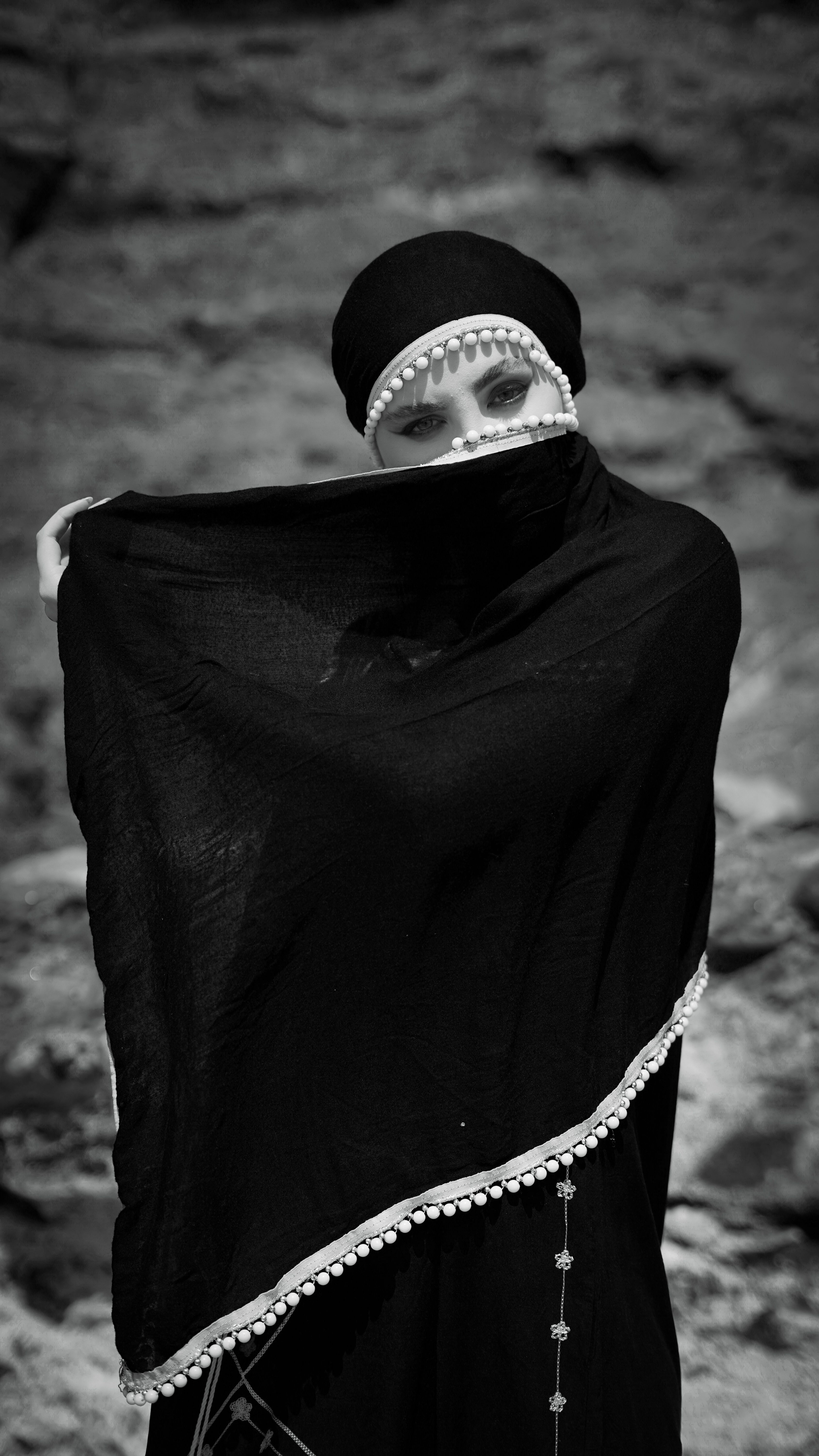 An enigmatic woman draped in a monochrome veil stands against a rocky outdoor background.