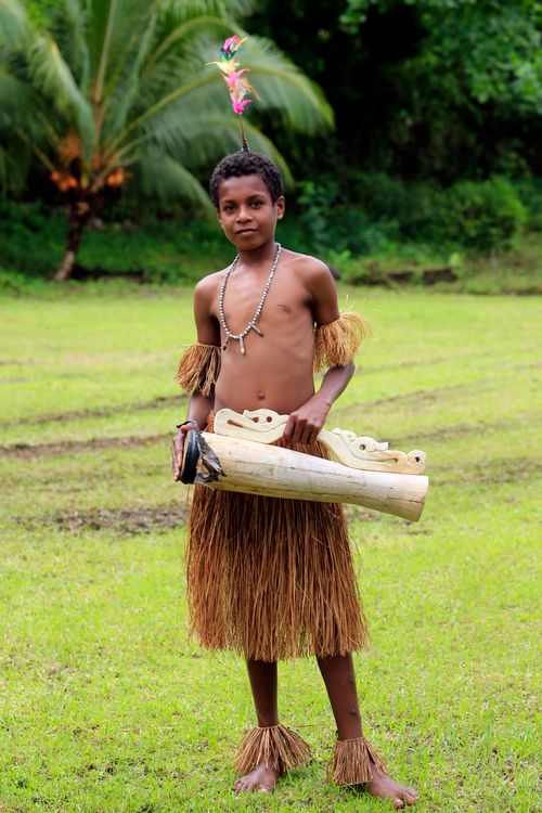 Free Young Papuan boy in traditional attire holding carved wooden items in a grassy outdoor setting. Stock Photo