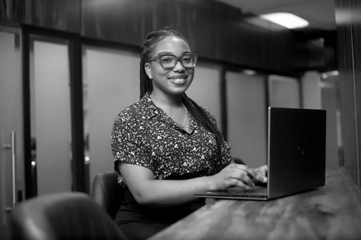 Black and white portrait of a smiling woman with braided hair using a laptop in an office.
