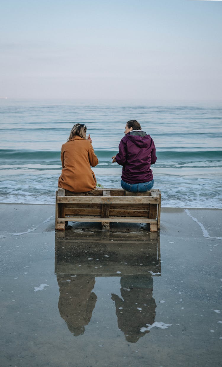 Two People Sitting On Pallet On Beach And Talking 