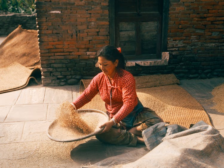 Woman Sitting On Straw Mat And Sifting Grain