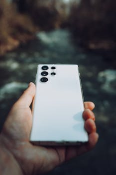 A close-up view of a hand holding a white smartphone outdoors, with a blurred background.