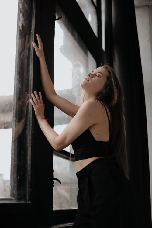 Free A woman in a black outfit poses elegantly by a large window indoors, capturing a serene moment. Stock Photo