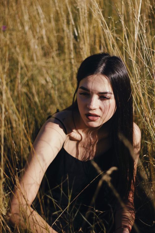 Free Portrait of a woman with long hair sitting in tall grass. Captured in a warm daylight setting. Stock Photo