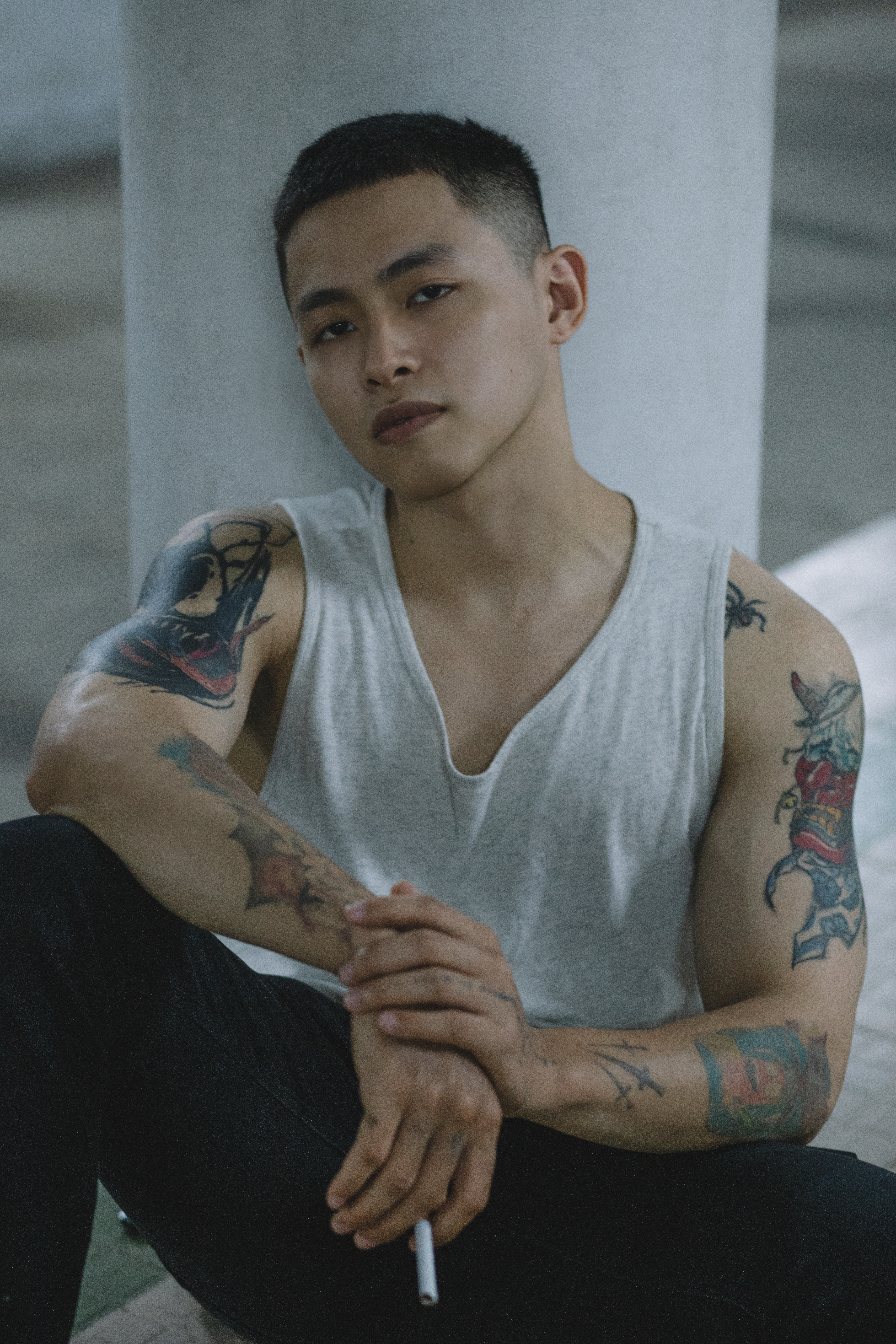A man with tattoos sits indoors, wearing a gray tank top, holding a cigarette.